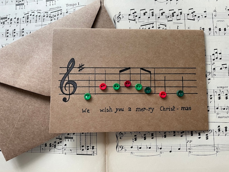 Może przedstawiać: Brązowa kartka świąteczna z motywem pięciolinii i napisem "we wish you a merry Christmas". Nuty muzyczne są reprezentowane przez czerwone i zielone guziki. Dołączona jest koperta.