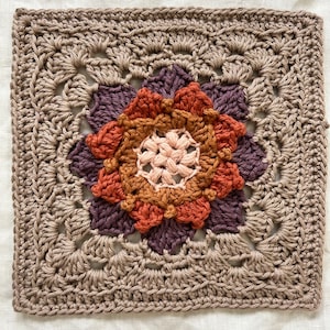May include: A crocheted square with a multi-colored flower design in the center. The flower is made of different shades of brown, orange, and purple yarn. The square is surrounded by a border of beige yarn.