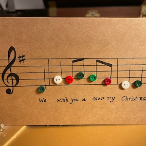 Może przedstawiać: Ręcznie robiona kartka świąteczna z nutami i napisem "We wish you a mer-ry Christmas". Nuty są reprezentowane przez kolorowe guziki w kolorze zielonym, czerwonym i białym, na brązowej kartce.
