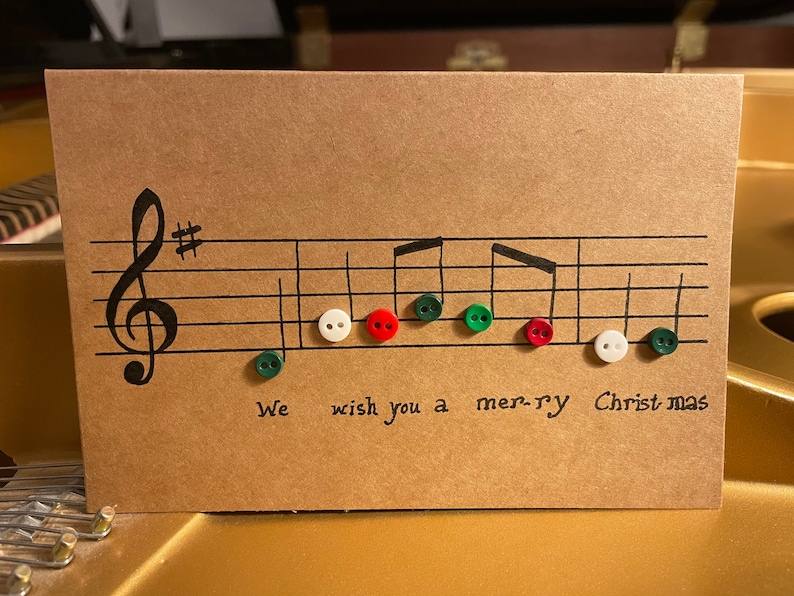 Może przedstawiać: Ręcznie robiona kartka świąteczna z nutami i napisem "We wish you a mer-ry Christmas". Nuty są reprezentowane przez kolorowe guziki w kolorze zielonym, czerwonym i białym, na brązowej kartce.