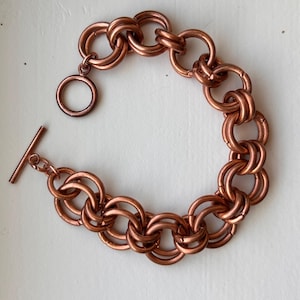May include: A copper-colored chainmail bracelet constructed from interlocking circular metal rings. The bracelet features a toggle clasp consisting of a circular ring and a straight bar, creating a continuous, repeating pattern of connected metallic links.