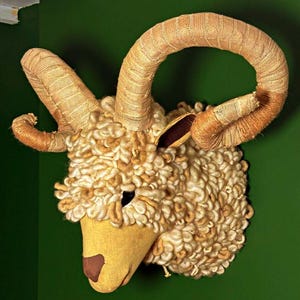 May include: A decorative ram head sculpture with large, curved horns. The horns are wrapped in a tan woven material, and the head features a soft, textured, off-white wool-like material. The ram's face is a light yellow colour with a brown nose and black eye.