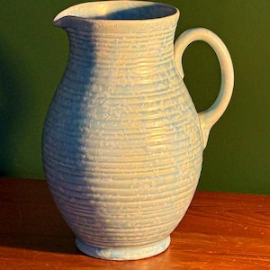 Puede incluir: Jarra de cerámica azul claro con un diseño acanalado horizontal texturizado. La jarra tiene un asa curva y un pico para verter. La base es de un tono azul más claro. La jarra está sobre una superficie de madera.