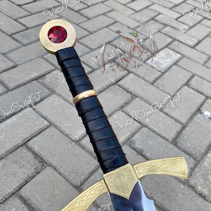 May include: A decorative sword with a black leather-wrapped handle and gold-coloured accents. The hilt features an ornate design, and the pommel has a red jewel. The blade is silver and reflects light.