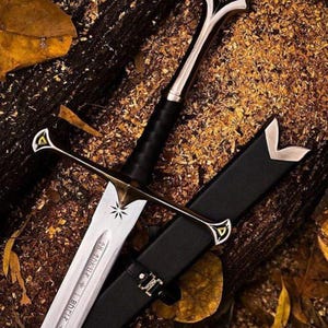 May include: A decorative sword with a silver blade and a black leather-wrapped handle. The sword has gold accents and a black sheath. The blade features etched text and a star design. The sword is displayed on a wooden surface.