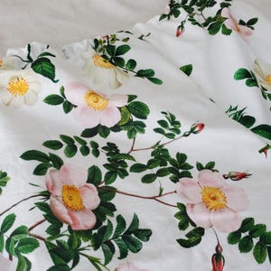 May include: A white fabric featuring a botanical print of light pink and white wild roses with yellow centres, green leaves, and small red buds. The material has a gathered, elasticated edge along the top, creating soft folds and drapes.