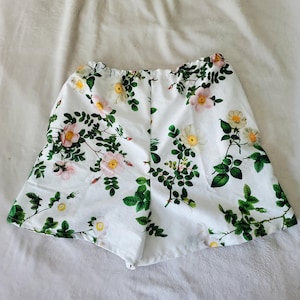 May include: White shorts featuring a botanical print of pink and white wild roses with green leaves and stems. The shorts have an elasticated waistband and a flared silhouette, showcasing a repeating floral pattern across the fabric.