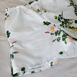 May include: White shorts featuring a botanical print of pink wild roses, green leaves, and small red buds. The garment has an elasticated waistband with gathered fabric detail, creating a soft, draped silhouette with a floral pattern throughout.