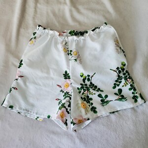 May include: White shorts featuring an elasticated waistband and a botanical print of pink and white wild roses with green leaves and stems. The garment has a loose, flared leg silhouette and is constructed from a lightweight, smooth fabric.