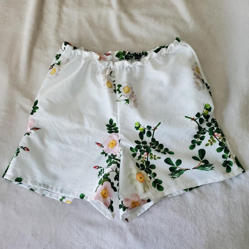 May include: White shorts featuring an elasticated waistband and a botanical print of pink and white wild roses with green leaves and stems. The garment has a loose, flared leg silhouette and is constructed from a lightweight, smooth fabric.