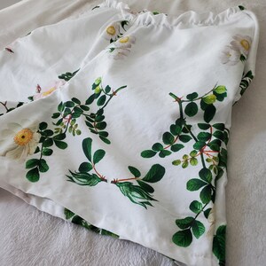 May include: White shorts featuring a botanical print of white flowers with yellow centres and green leaves on stems. The shorts have a gathered, elasticated waistband at the top, creating a ruffled edge. The fabric is white with a floral pattern.