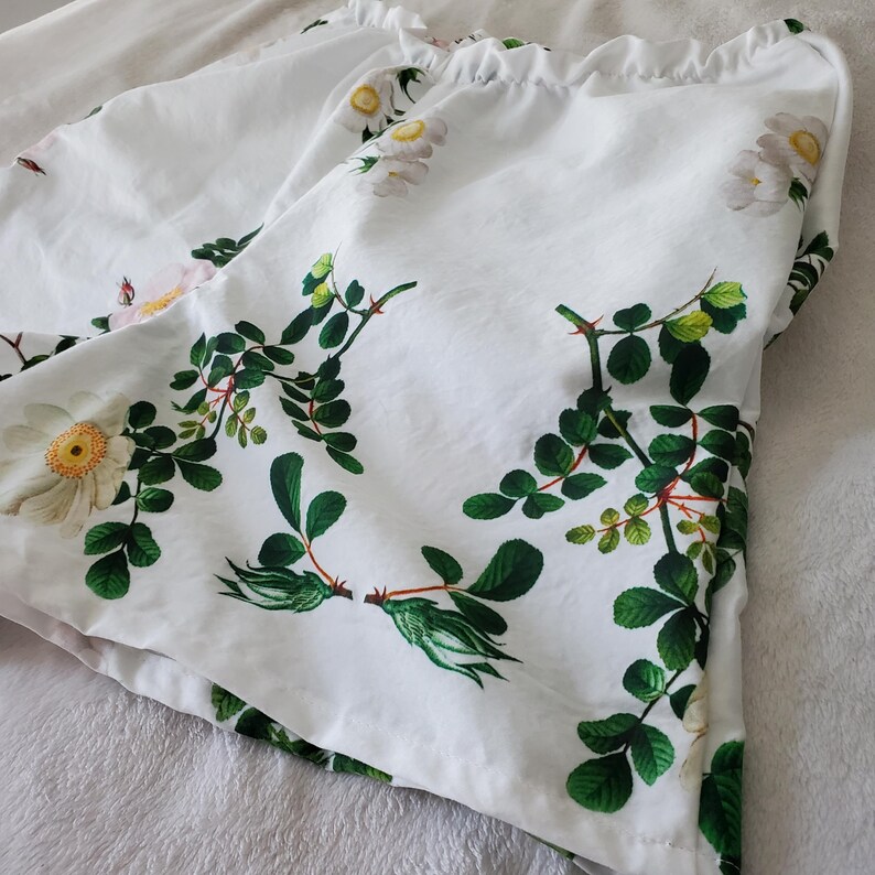 May include: White shorts featuring a botanical print of white flowers with yellow centres and green leaves on stems. The shorts have a gathered, elasticated waistband at the top, creating a ruffled edge. The fabric is white with a floral pattern.