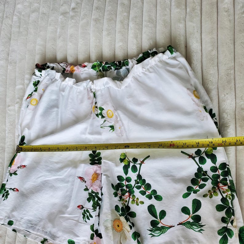 May include: White shorts featuring a floral pattern of pink flowers, yellow centres, and green leaves. A yellow measuring tape is placed across the front, showing a width of approximately 51 cm across the waistband area.