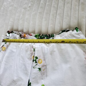 May include: White shorts featuring a floral pattern of pink and white flowers with green leaves. A yellow measuring tape is placed across the waistband, showing a width of approximately 38 cm. The garment has an elasticated waistband.
