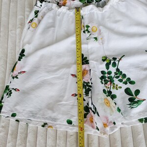 May include: White shorts featuring a floral pattern of pink and white roses with green leaves. A yellow measuring tape is placed vertically over the centre, showing a length of approximately 40.5 centimetres from the waistband to the bottom hem.