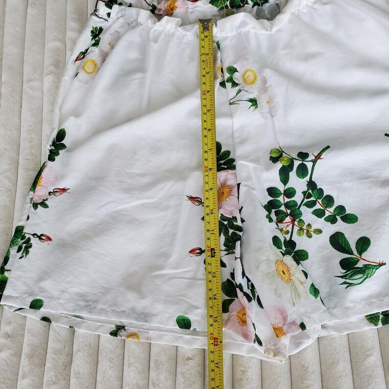 May include: White shorts featuring a floral pattern of pink and white roses with green leaves. A yellow measuring tape is placed vertically over the centre, showing a length of approximately 40.5 centimetres from the waistband to the bottom hem.