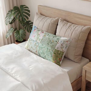 Peut inclure: Un lit avec une couette blanche et deux oreillers beiges matelassés. Un long oreiller rectangulaire avec un motif patchwork de motifs floraux dans les tons de vert, rose et bleu repose sur le lit.