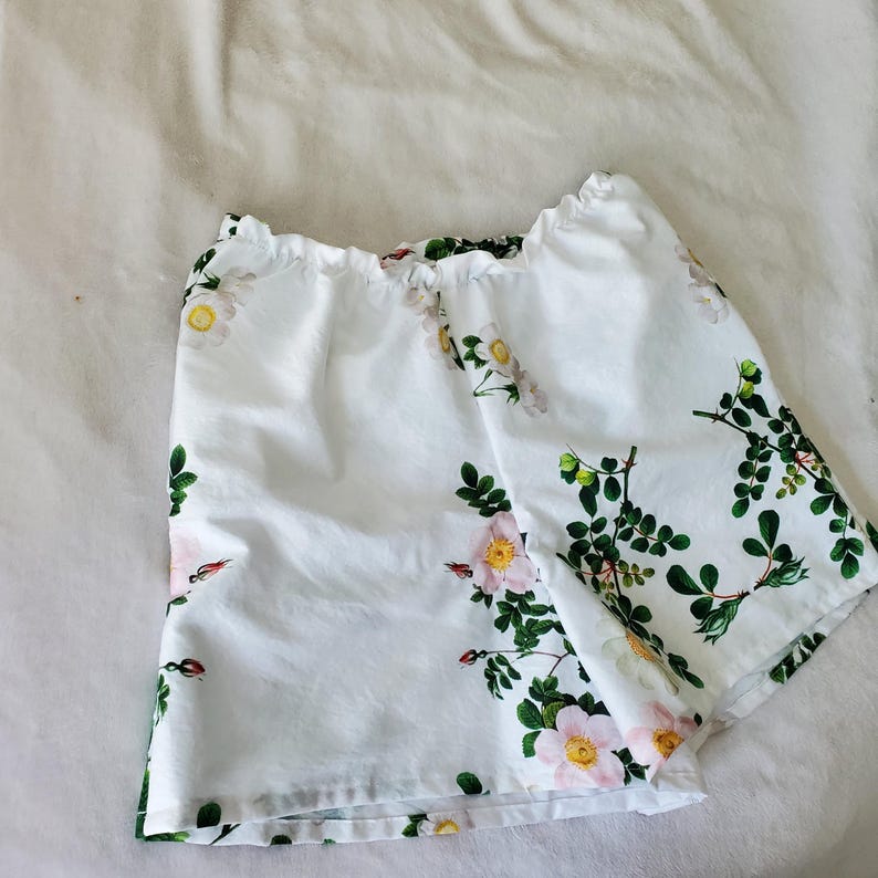 May include: White shorts featuring a botanical print of pink and white wild roses with green leaves and stems. The shorts have an elasticated, gathered waistband and a loose, relaxed fit, showcasing a floral pattern across the fabric.