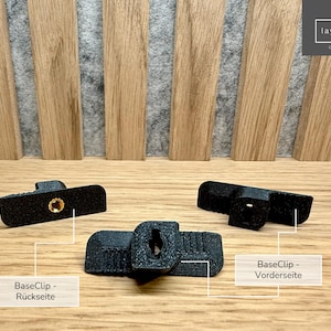 May include: Three black plastic clips, two of which are stacked on top of each other. The clips have a small hole in the centre and a textured surface. The text "BaseClip - Vorderseite" is visible in the image. The text "BaseClip - Rückseite" is also visible in the image.
