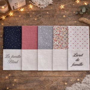 Puede incluir: Cinco fundas rectangulares de tela dispuestas una al lado de la otra. La funda izquierda es azul oscuro con estrellas doradas y el texto La famille Blind. Las otras presentan rosa sólido, gris con estrellas blancas, motivos florales y corazones rojos con Livret de famille.