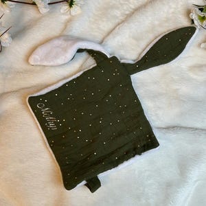 May include: A square olive green security blanket with a white plush backing and two long rabbit ears. The green fabric features a gold star pattern and the embroidered name "Marilyn" in silver thread on the front corner.