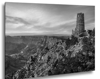 May include: Black and white photograph of a scenic landscape featuring a deep canyon and a stone tower. The tower is perched on a cliff edge, overlooking the vast canyon with a river winding through it. The sky is filled with clouds.