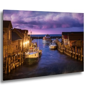 Könnte beinhalten: Ein malerischer Druck eines Hafens in der Abenddämmerung. Holzhäuser mit warmer Beleuchtung säumen das Ufer. Mehrere Boote liegen im Hafen vor Anker, mit einem leuchtenden lila und rosa Himmel darüber. Das Wasser ist tiefblau.