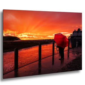 Könnte beinhalten: Ein Foto zeigt eine Person mit einem roten Regenschirm auf einem nassen Steg. Der Himmel leuchtet in Orange- und Rottönen während eines dramatischen Sonnenuntergangs. Im Hintergrund ist ein Boot zu sehen, und die Holzpfosten des Stegs spiegeln sich im Wasser.