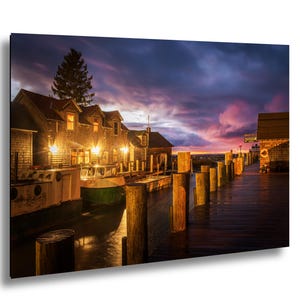 Könnte beinhalten: Ein malerischer Druck eines Hafens in der Abenddämmerung. Holzhäuser mit beleuchteten Fenstern säumen das Ufer. Ein grünes und weißes Boot liegt im Dock. Der Himmel ist eine Mischung aus Lila, Orange und Blau.