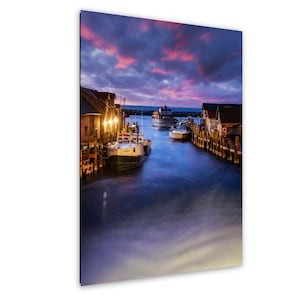 Könnte beinhalten: Ein malerischer Druck eines Hafens in der Abenddämmerung. Das Bild zeigt Boote, die an Holzstegen angedockt sind, mit Gebäuden, die von warmen Lichtern beleuchtet werden. Der Himmel ist mit leuchtend rosa und lila Wolken gefüllt, die sich im ruhigen Wasser spiegeln.