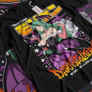 Puede incluir: Camiseta negra con una ilustración colorida de un personaje femenino con cabello verde y ropa morada. El texto "Morrigan" y "Darkstalkers" es visible en la camiseta.