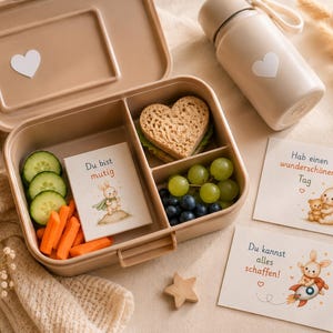 Könnte beinhalten: Eine beige Lunchbox mit einem weißen Herzaufkleber, gefüllt mit einem herzförmigen Sandwich, Trauben, Blaubeeren, Gurkenscheiben und Karottensticks. Eine passende Wasserflasche und Karten mit Hasenillustrationen sind ebenfalls vorhanden.