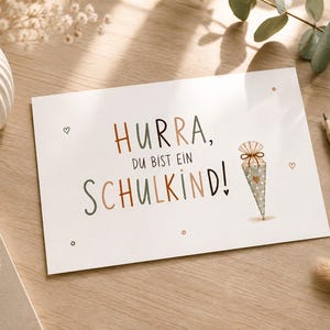 Puede incluir: Una tarjeta de felicitación rectangular blanca con el texto "HURRA, DU BIST EIN SCHULKIND!" en letras de color marrón y gris oscuro. A la derecha del texto hay una ilustración de un pequeño cono con lunares y un lazo.