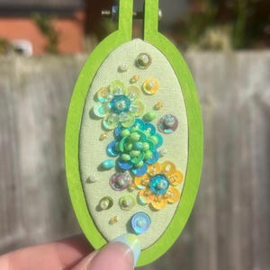 May include: A small, oval-shaped embroidery hoop with a light green fabric background. The hoop is framed in bright green wood. The fabric is decorated with three sequined flowers in shades of blue, yellow, and green, with small pearl beads.