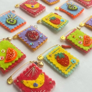 Pode incluir: Uma coleção de broches de feltro coloridos com desenhos de frutas, incluindo morangos, bananas e kiwis. Cada broche quadrado tem um alfinete de segurança dourado anexado. Os broches são em várias cores como vermelho, amarelo, verde e azul.