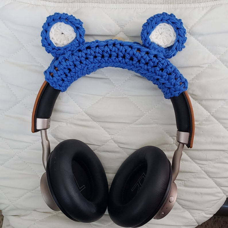K&ouml;nnte beinhalten: Blaue geh&auml;kelte Kopfh&ouml;rerabdeckung mit wei&szlig;en ohrenf&ouml;rmigen Akzenten. Die Abdeckung befindet sich auf einem Paar schwarzer Over-Ear-Kopfh&ouml;rer.