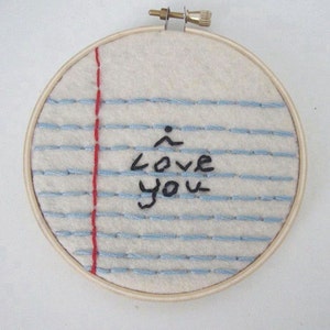 Könnte beinhalten: Ein weißer Stickrahmen mit blauen und roten Stichen, die einen Notizbuch-Papier-Effekt erzeugen. Die Worte "Ich liebe dich" sind in schwarzem Faden gestickt.