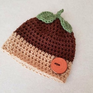 May include: A brown and tan crocheted hat with a green crocheted leaf on top and a brown button. The hat is designed to look like a pudding.