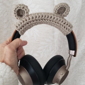 May include: Beige crocheted headband with bear ears, covering the headband of a pair of black over-ear headphones with brown accents.
