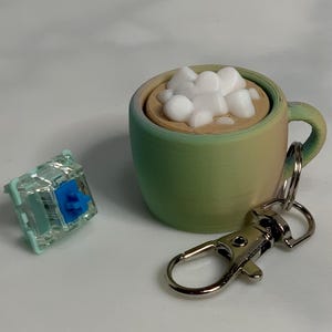 Może przedstawiać: Zielony i niebieski kubek ceramiczny z białym uchwytem, wypełniony białymi piankami i brązowym płynem. Kubek jest przymocowany do srebrnego breloczka.