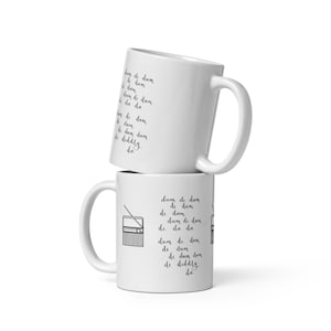 May include: Two white ceramic mugs stacked on top of each other. The top mug has the phrase "dum de dum de dum de dum da da" written in black script. The bottom mug has the same phrase and a drawing of a radio.
