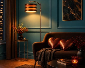May include: A floor lamp with a dark metal stand and three stacked cylindrical shades emitting a warm glow. The lamp stands beside a dark sofa featuring two heart-shaped sequined pillows and a gray throw blanket, with a side table nearby.