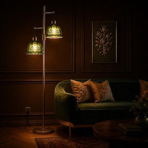May include: A two-light floor lamp with green glass shades and a gold-colored metal frame. The lamp is positioned in a room with a dark green velvet sofa, gold pillows, and a dark brown wall with a framed floral artwork.