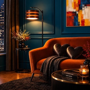 May include: A burnt orange velvet sofa featuring two heart-shaped sequined pillows and a dark gray throw blanket. Beside the sofa stands a black metal floor lamp with a tiered wooden shade and a small round side table holding a floral arrangement.