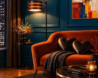 May include: A burnt orange velvet sofa featuring two heart-shaped sequined pillows and a dark gray throw blanket. Beside the sofa stands a black metal floor lamp with a tiered wooden shade and a small round side table holding a floral arrangement.