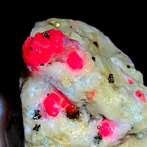 Puede incluir: Un espécimen mineral rugoso de color beige que presenta múltiples inclusiones fluorescentes de color rosa neón vibrante y pequeñas motas metálicas oscuras dispersas por su superficie texturizada e irregular bajo luz ultravioleta.