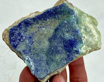 May include: A rough-cut stone with a striking combination of colors. The stone features a deep blue center, transitioning to lighter blue and green hues towards the edges. The outer edges are a light tan color, creating a natural, earthy appearance.