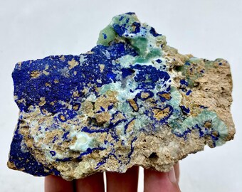 May include: A natural mineral specimen featuring a striking combination of colors. Deep blue azurite crystals are interspersed with patches of light green malachite and set against a beige matrix. The rough, textured surface showcases the raw beauty of the stone.