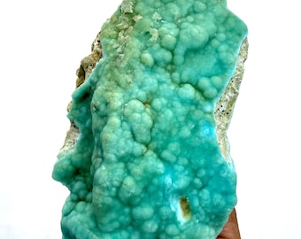 May include: A large, irregular piece of teal-colored mineral with a bubbly, textured surface. The mineral has a rough, beige-colored edge and a few small holes. The overall shape is organic and natural, with varying shades of teal.
