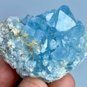 May include: A cluster of translucent light blue celestite crystals with sharp, geometric facets. The mineral specimen features a rough, textured base with white and grey mineral inclusions, contrasting with the clear, bright blue crystalline points.